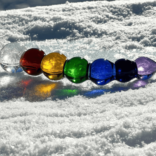 Photographie dans la neige d’un dildo sur fond blanc, il représente un arc-en-ciel, il est coloré de blanc, rouge, jaune, vert, bleu, marine, violet, un festival de confettis et de couleurs
