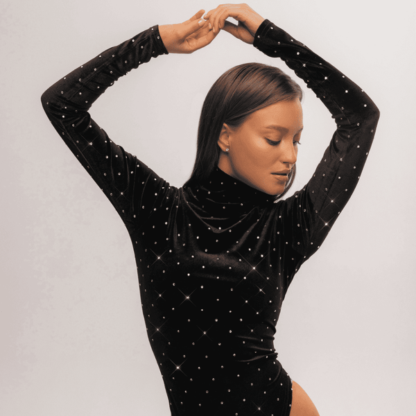 Une femme se tient élégamment devant un mur blanc, vêtue d'un body en velours noir au col roulé. Ce vêtement raffiné est rehaussé par l'incrustation minutieuse de cristaux Swarovski, ajoutant une touche de brillance et d'éclat à sa tenue. L'association du velours sombre avec les cristaux scintillants crée un contraste visuel captivant, mettant en valeur la sophistication et le souci du détail dans le choix de sa garde-robe. Cette combinaison unique de textures et d'éléments décoratifs confère à l'ensemble u