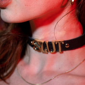 Gros plan sur un choker en cuir noir LIEBE SEELE avec lettres métalliques dorées formant le mot SLUT, porté autour du cou, éclairage rouge et ambiance studio