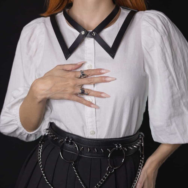Choker 86647 en cuir noir, porté avec une chemise blanche et une jupe noire à détails argentés. Confectionné à la main, avec des vis à l’avant rappelant les boutons d’un col et une boucle réglable à l’arrière.