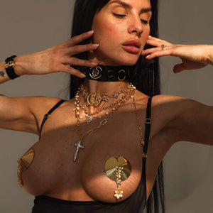 Femme portant plusieurs colliers dorés QRUCIFIX et des nipple covers métalliques en forme de disque avec pendentifs. Elle pose avec les bras relevés, yeux fermés. Choker noir avec logo et bretelles de lingerie noire visibles.