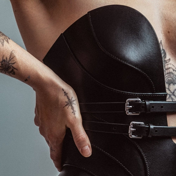 Mannequin portant le corset Daelos en cuir noir RE:STRICT chez Brigade Mondaine Paris, décolleté plongeant structuré, sangles ajustables, porté avec accessoires assortis sur fond rouge.