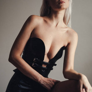 Mannequin portant le corset Daelos en cuir noir RE:STRICT chez Brigade Mondaine Paris, décolleté plongeant structuré, sangles ajustables, porté avec accessoires assortis sur fond rouge.