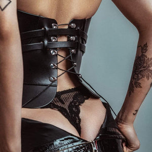 Mannequin portant le corset Daelos en cuir noir RE:STRICT chez Brigade Mondaine Paris, décolleté plongeant structuré, sangles ajustables, porté avec accessoires assortis sur fond rouge.