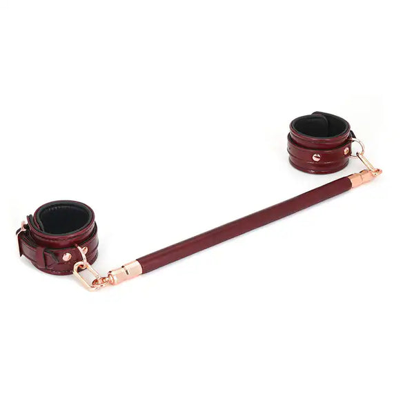 Barre d’écartement en cuir rouge vin avec noyau en bois d’eucalyptus. Joints toriques en métal or rose pour fixer des chevillières. Élégante et fonctionnelle pour des jeux BDSM raffinés.
