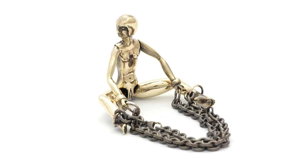 Ceci est un cockring en forme de poupée en bronze patiné. Tout en tournant la tête du côté droit, elle écarte les jambes et elle a des chaînes aux pieds.