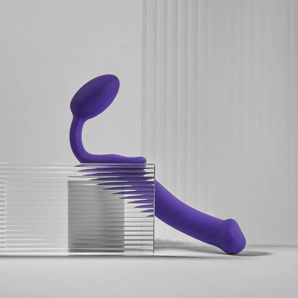 Plug strapless violet STRAP-ON-ME au design courbé et ergonomique, posé sur un socle minimaliste, sélection Brigade Mondaine Paris.