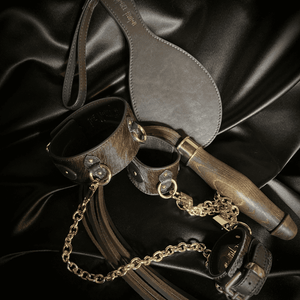 Photographie sur fond de textile noir, accessoires bdsm collier, laisse, menottes et fouet de cuir marron et détails dorés comme chaines et cadenas
