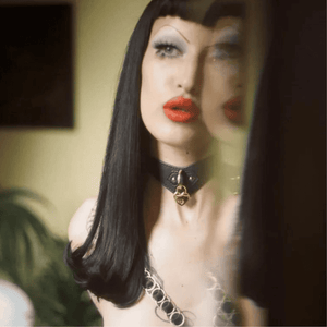 Photographie prise dans le reflet d’un miroir, une femme brune avec une frange courte, des soursiles fins et maquillage porte un collier de cuir noir avec cadenas coeur