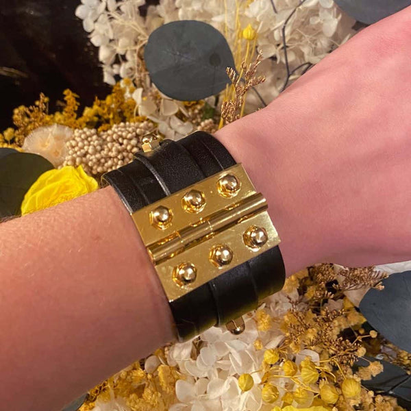 Bracelet à charnière à double sangle en cuir noir, ajustable de 17 à 20 cm. Confectionné à la main par Úna Burke en Irlande, avec cuir de vachette pleine fleur et laiton plaqué or. Design rock chic et durable.