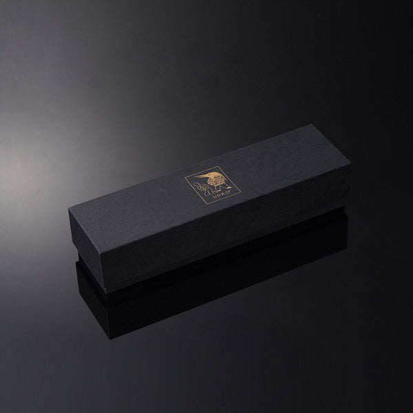 Le packaging UPKO, noir et doré, est disposé en diagonale sur un fond noir brillant qui capte et reflète la lumière blanche.