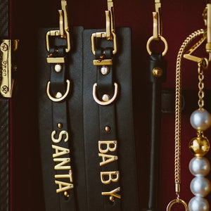 Accessoires UPKO en cuir noir avec elastiques réglables, perles nacrées et finitions métalliques dorées, comprenant colliers, menottes et pièces personnalisées, présentés sur fond bordeaux, disponibles chez Brigade Mondaine Paris.