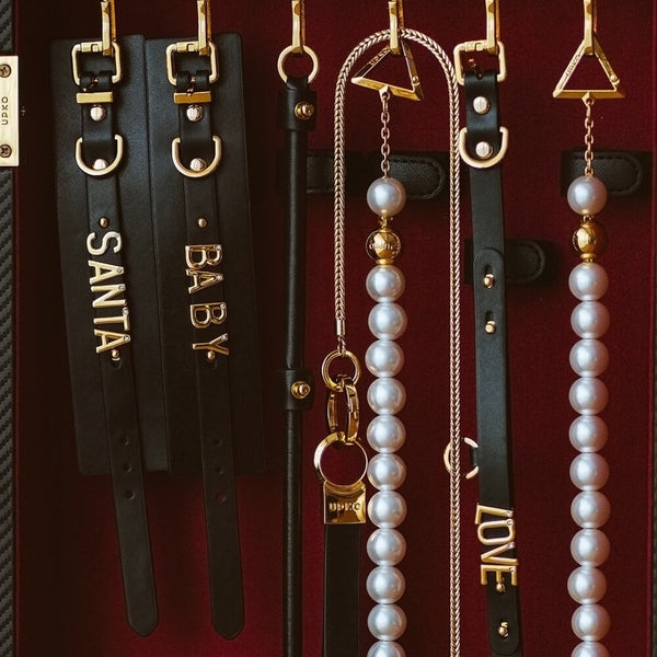 Accessoires UPKO en cuir noir avec elastiques réglables, perles nacrées et finitions métalliques dorées, comprenant colliers, menottes et pièces personnalisées, présentés sur fond bordeaux, disponibles chez Brigade Mondaine Paris.