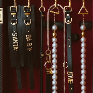 Accessoires UPKO en cuir noir avec elastiques réglables, perles nacrées et finitions métalliques dorées, comprenant colliers, menottes et pièces personnalisées, présentés sur fond bordeaux, disponibles chez Brigade Mondaine Paris.