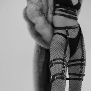 Dans une ambiance en noir et blanc, une femme porte le Harnais CLASSIC 3 en cuir – ST. VENDETTA, mettant en valeur sa silhouette avec élégance. Sous un manteau en fourrure luxueux, les jarretières Gorgona en cuir accentuent ses jambes, apportant une touche de sensualité et de raffinement à son look audacieux.