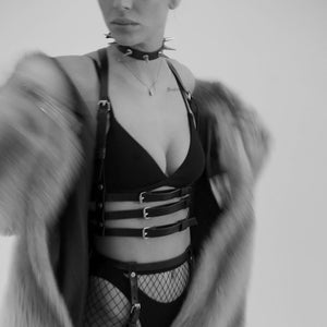 Femme en noir et blanc portant un harnais en cuir naturel fait à la main, avec trois lanières sous le buste et deux lanières montant vers le cou, créant un effet de soutien-gorge. Elle porte également un soutien-gorge noir, et le cuir est mis en valeur par la lumière, accentuant la texture et le design sophistiqué.