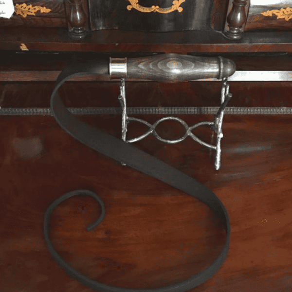 Photographie d’un fouet avec un manche en bois de la forme d’un penis avec fouet en cuir noir, l’objet est posé sur un piédestal de fer sur un parquet aux couleurs chaudes