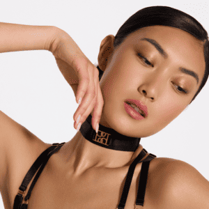 Photograph of a woman with a black chocker and Bordelle logo in gold on the front, she has a make-up-age your legends and natural and her hair plated and tied up.