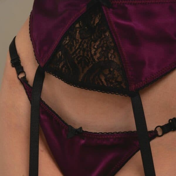 Detail einer Frau in 3/4-Länge, die einen Triangel-Bralette-BH, einen Strapsgürtel und ein offenes Höschen aus violettem Satin und schwarzer Spitze trägt.